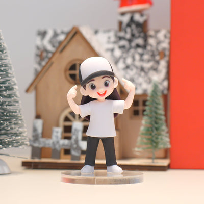 Figurine of a person in a white shirt and black pants with a white cap, standing in front of a decorative background with trees and a house.