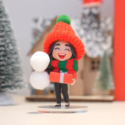 Decorative figurine of a person wearing a red hat and scarf, holding snowballs and a gift, with a festive background.