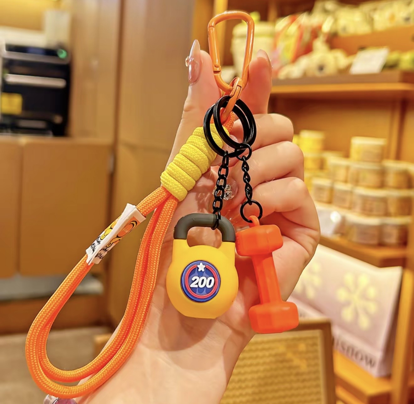 Hand holding a keychain with a yellow lock, black carabiner, and orange dumbbell charm in a store setting.