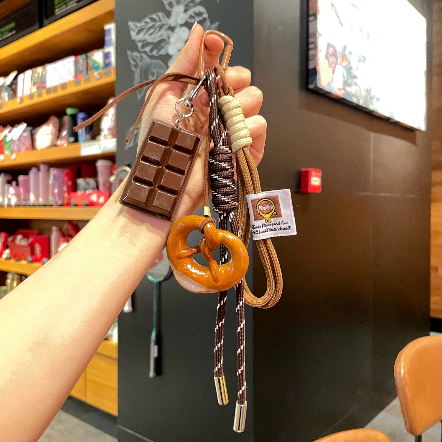 Hand holding a keychain with chocolate bar, pretzel, and rope design in a store setting.