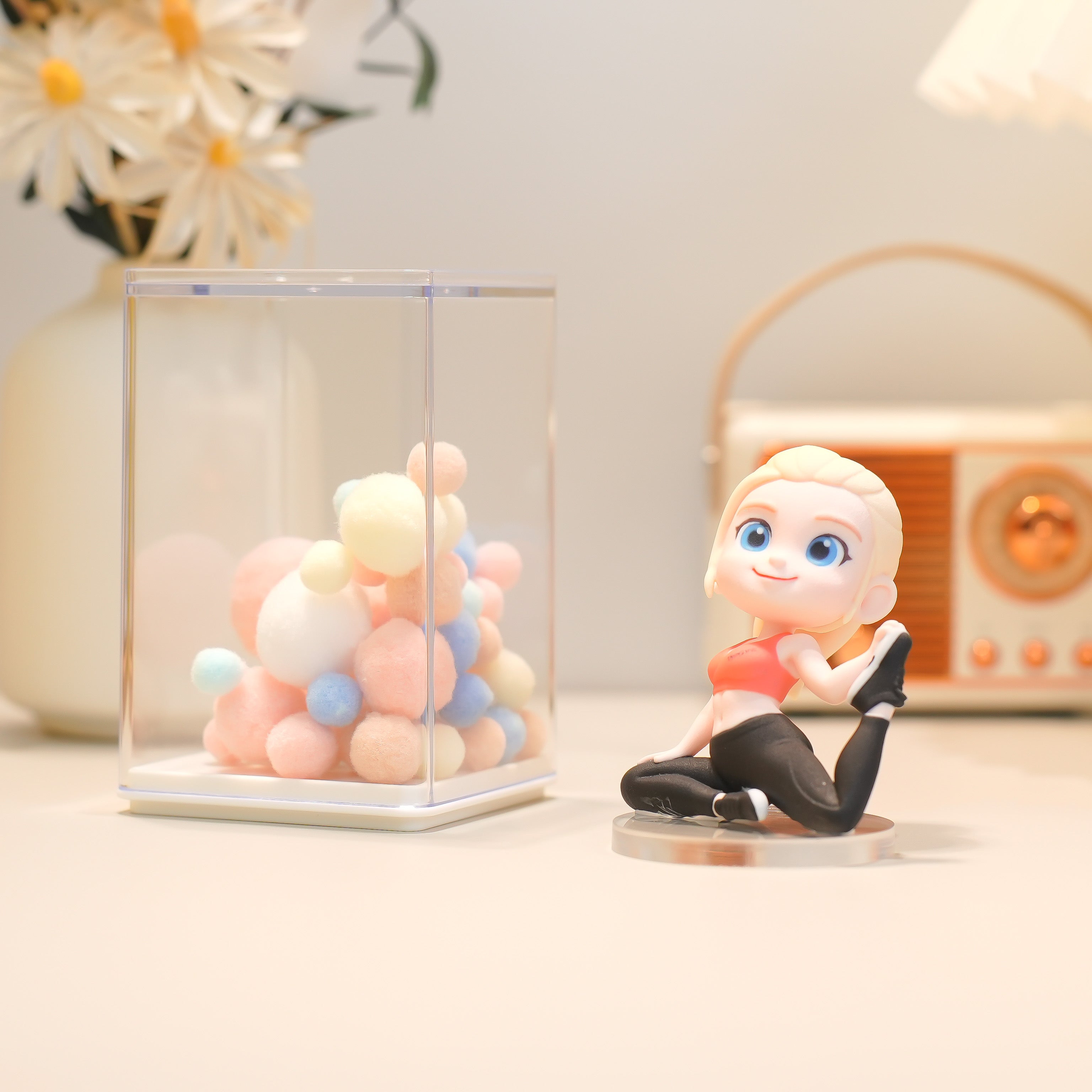 Decorative figurine of a blonde woman sitting next to a glass cube with colorful balloons on a light surface.