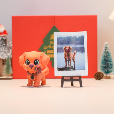 Small orange dog figurine next to a photo of a dog on a lake with a red box and Christmas tree in the background.