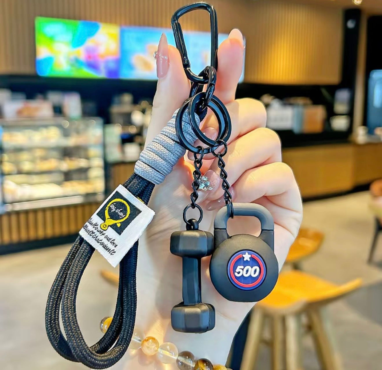 Hand holding a keychain with a dumbbell and kettlebell design, labeled '500', in a blurred indoor setting.