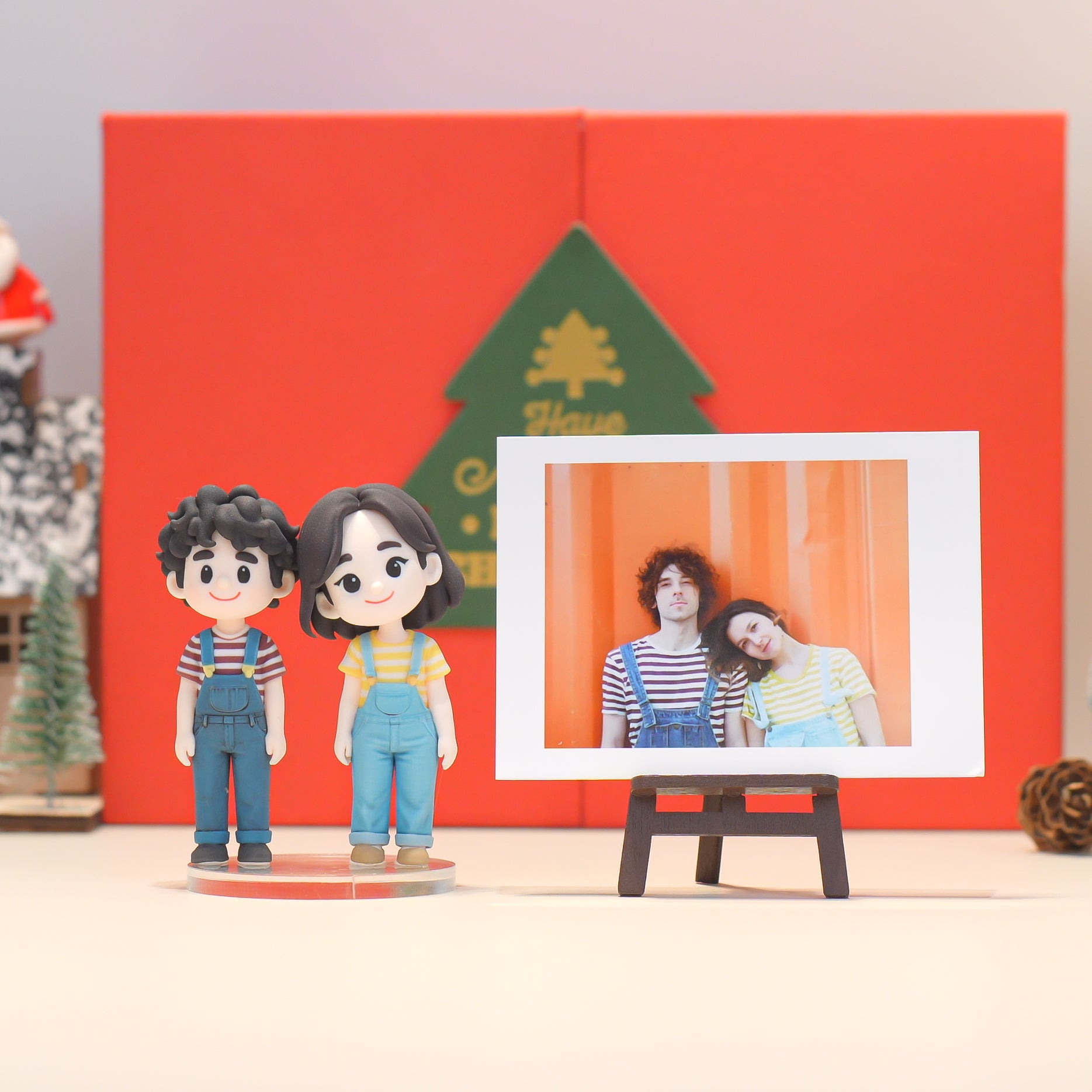 Two figurines of a couple in front of a framed photo on a red background with a Christmas tree.