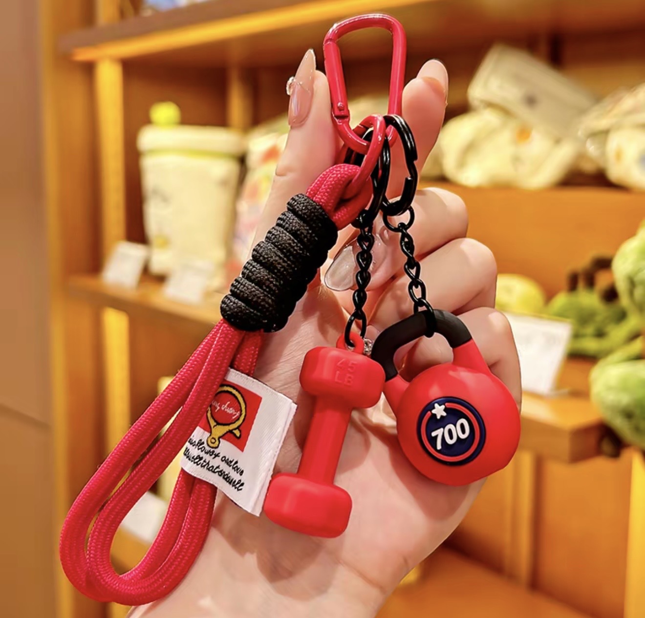 Red keychain with dumbbell-shaped ends held by a hand in front of a blurred store shelf.