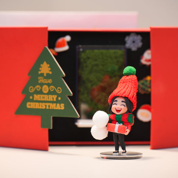 Figurine of a person in winter clothing holding a snowball and gift, with a 'Merry Christmas' tree decoration.